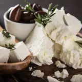 Feta, un formaggio ricco di proteine per 100g, servito con olive nere e rametti di rosmarino su un tagliere di legno.