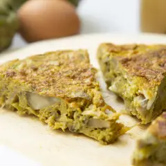 Fette di frittata con pecorino e verdure su tagliere di legno, con uova e carciofi sullo sfondo.