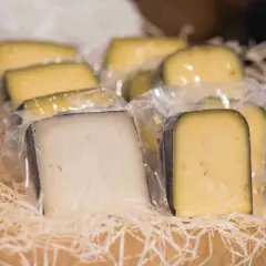 Vari tipi di formaggio fuori dal frigo, confezionati singolarmente e disposti su paglia in una cesta di legno.