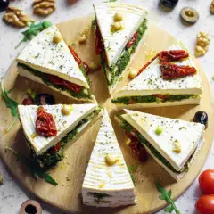 Fette di primo sale al forno, farcite con pesto e pomodori secchi, disposte a raggiera su un tagliere, con noci e pomodorini intorno.