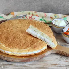 Torta salata con formaggi avanzati, soffice e dorata, con una fetta tagliata che rivela il ripieno cremoso. Accanto, ciotoline con salse e verdure sottaceto.