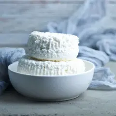Due forme di ricotta magra impilate in una ciotola bianca, con un drappo grigio sullo sfondo.