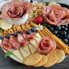 Presentazione tagliere aperitivo con salumi a rosa, formaggi, mirtilli, fragole, pretzel e grissini.