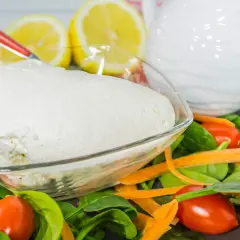 Un fresco quark formaggio in una ciotola, circondato da spinaci, carote e pomodorini. Limoni e una brocca bianca sullo sfondo.