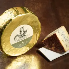 Formaggio blu con crosta scura, accanto a una forma intera avvolta in carta dorata. Questo cacio cos'è? Un'eccellenza da gustare.