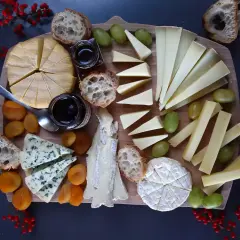 Un ricco tagliere con vari formaggi da aperitivo, pane croccante, uva, albicocche secche e marmellata.