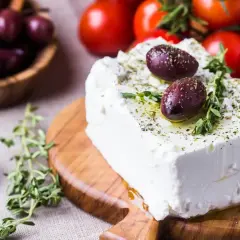 Un blocco di feta greca condita con erbe aromatiche e olive su un tagliere di legno, con pomodori e olive in sottofondo.