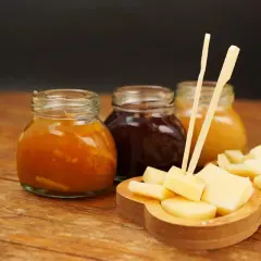 Tre vasetti di marmellata, uno arancione, uno scuro e uno giallo, accompagnano un tagliere di formaggio a cubetti, pronto per essere gustato.