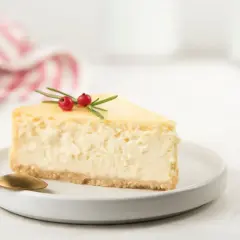 Una fetta di cheesecake con yogurt greco, guarnita con rosmarino e bacche rosse, su un piatto bianco con un cucchiaino dorato.
