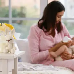 Mamma nutre il neonato con il biberon. Sul tavolino, un coniglietto di peluche e accessori per la pappa, come i latticini e derivati.