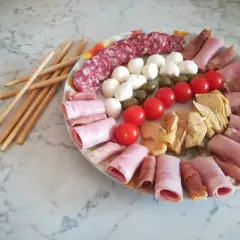 Un'idea per un tagliere di salumi: salame, mozzarelline, olive, pomodorini, carciofini e involtini di prosciutto cotto e crudo, con grissini a lato.