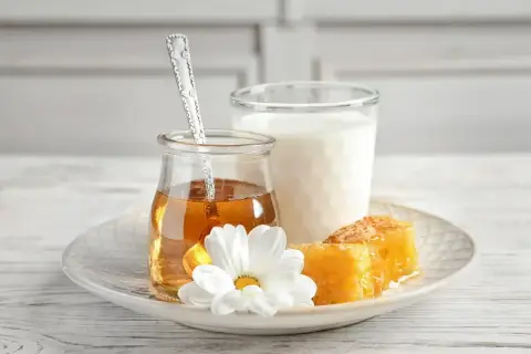 Un bicchiere di latte, un vasetto di miele dorato con un cucchiaino, e favi di miele su un piatto con un fiore bianco. Alcuni credono che latte caldo e miele faccia male.