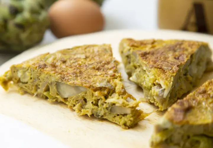 Fette di frittata con pecorino e verdure su tagliere di legno, con uova e carciofi sullo sfondo.