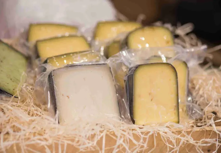 Vari tipi di formaggio fuori dal frigo, confezionati singolarmente e disposti su paglia in una cesta di legno.