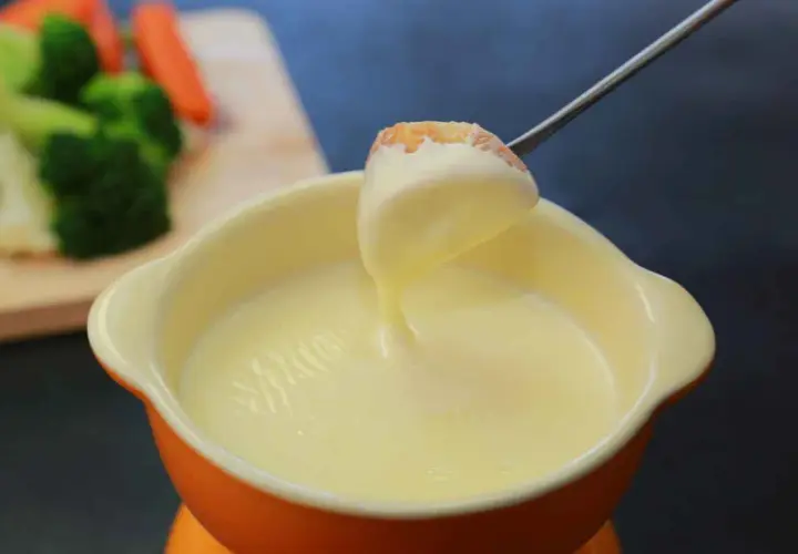 Una forchetta intinge un pezzo di pane in una cremosa fonduta per pasta. Sullo sfondo, verdure fresche.