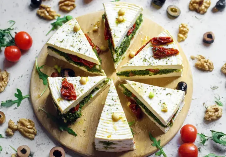 Fette di primo sale al forno, farcite con pesto e pomodori secchi, disposte a raggiera su un tagliere, con noci e pomodorini intorno.