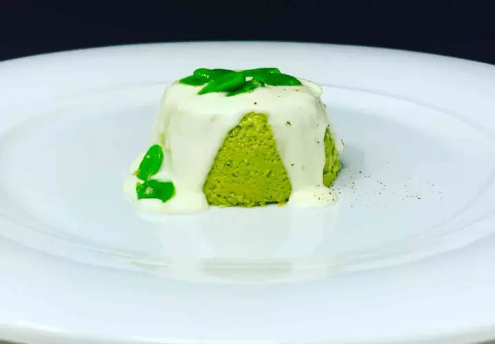 Sformato di pecorino verde brillante, ricoperto da una salsa bianca cremosa e guarnito con foglie verdi fresche.