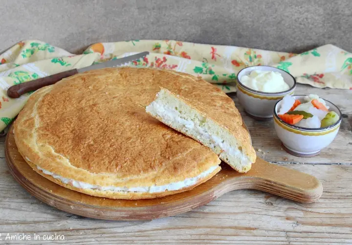 Torta salata con formaggi avanzati, soffice e dorata, con una fetta tagliata che rivela il ripieno cremoso. Accanto, ciotoline con salse e verdure sottaceto.