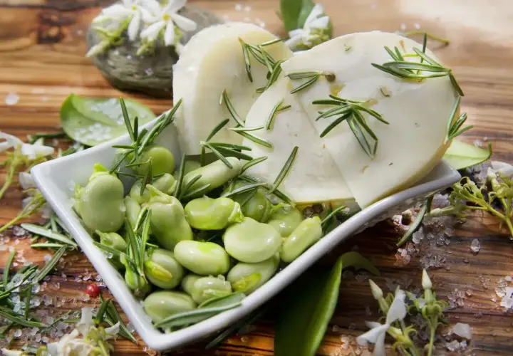 Baccelli freschi e fette di pecorino guarnite con rosmarino su un tagliere di legno.