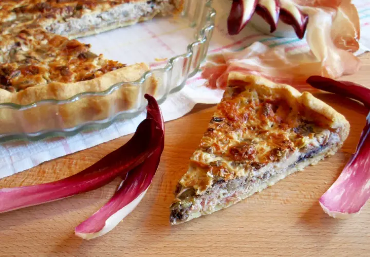 Una fetta di torta salata con taleggio, ricca e dorata, accanto a foglie di radicchio e prosciutto crudo.