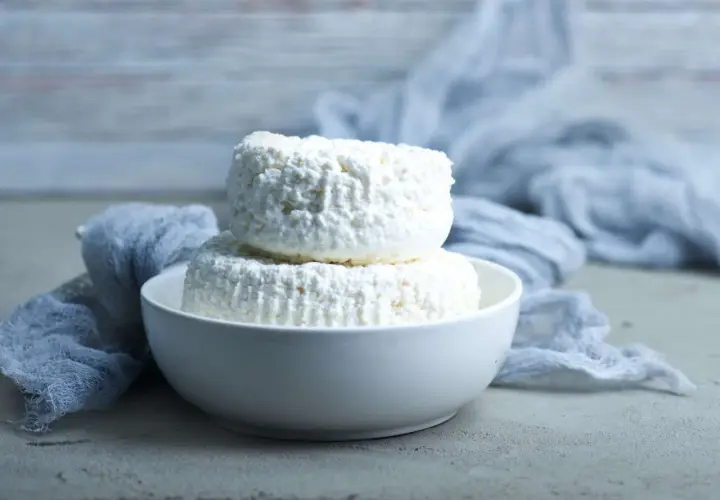 Due forme di ricotta magra impilate in una ciotola bianca, con un drappo grigio sullo sfondo.