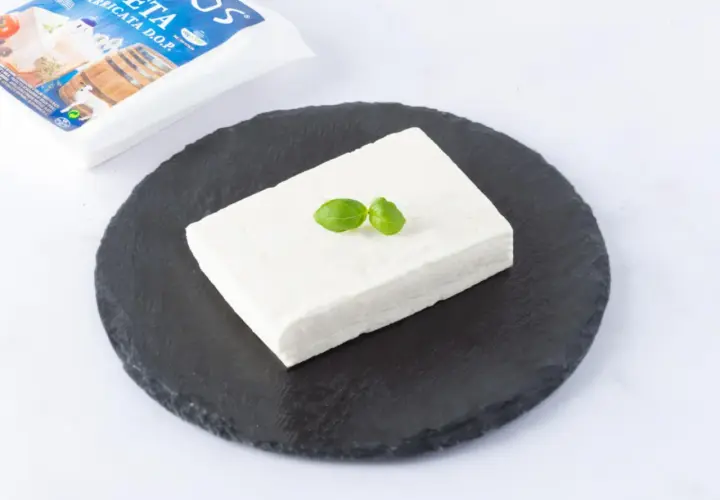 Un blocco di feta DOP su una pietra scura, guarnito con basilico. La confezione è visibile sullo sfondo.