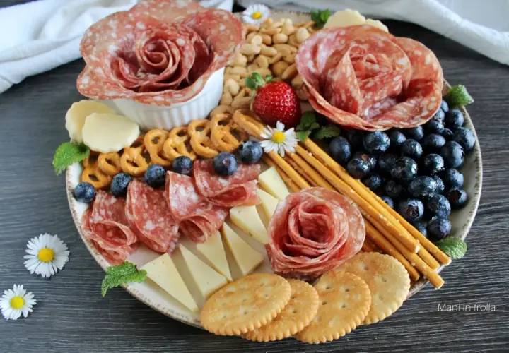 Presentazione tagliere aperitivo con salumi a rosa, formaggi, mirtilli, fragole, pretzel e grissini.