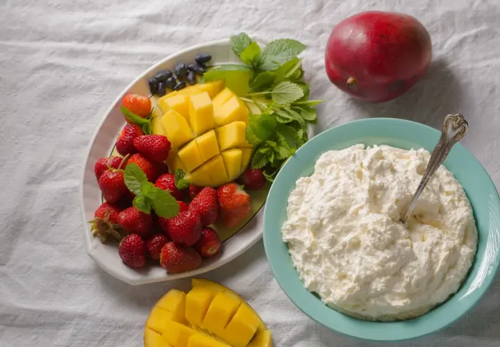 Un piatto di fragole, mango e mirtilli accanto a una ciotola di cosa sono i fiocchi di latte, con un mango intero sullo sfondo.