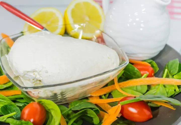 Un fresco quark formaggio in una ciotola, circondato da spinaci, carote e pomodorini. Limoni e una brocca bianca sullo sfondo.