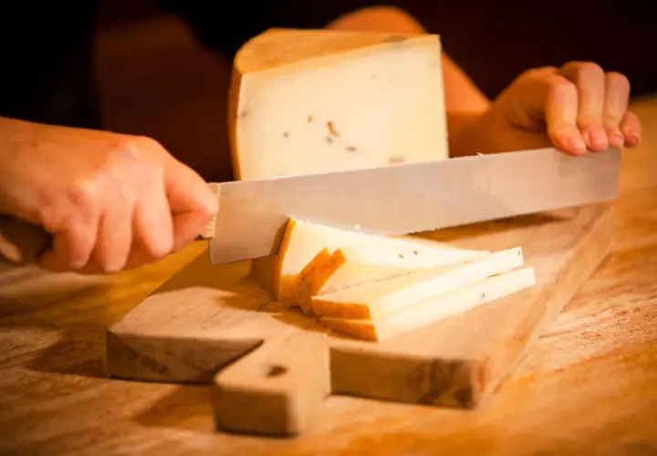 Mani che tagliano formaggio su un tagliere di legno. Fette sottili e un pezzo più grande di formaggio stagionato.