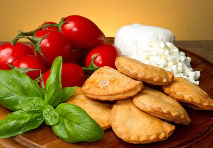 Panzerotti pugliesi dorati, pomodorini freschi, mozzarella a cubetti e basilico profumato su un tagliere di legno.