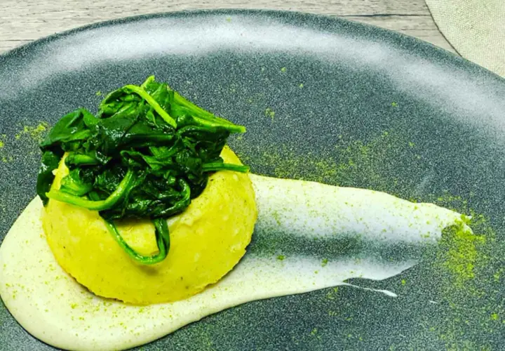 Flan di pecorino giallo, sormontato da spinaci freschi, su una crema bianca e polvere verde su piatto scuro.