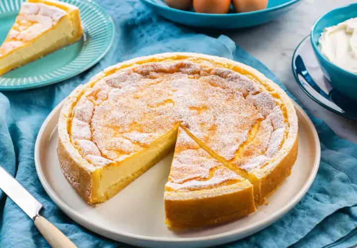 Torta soffice e dorata, simile a un quark formaggio, cosparsa di zucchero a velo. Una fetta è già servita.