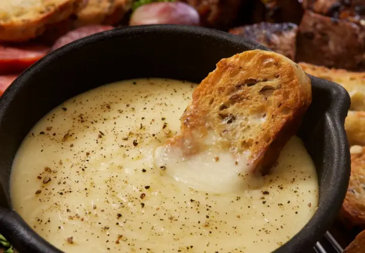 Una fetta di pane croccante si tuffa in una cremosa fonduta di formaggio, cosparsa di pepe nero. Sullo sfondo, salumi e pane tostato invitano a un convivio.