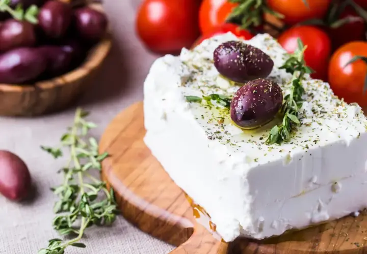 Un blocco di feta greca condita con erbe aromatiche e olive su un tagliere di legno, con pomodori e olive in sottofondo.