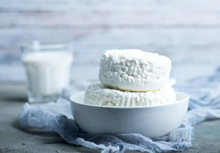 Due forme di ricotta fresca in una ciotola bianca, con un bicchiere di latte sullo sfondo. La ricotta si può congelare per conservarla più a lungo.