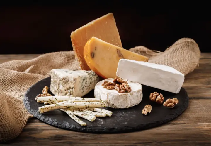 Selezione di formaggi grassi: un pezzo di formaggio erborinato, un brie, un formaggio stagionato e bastoncini di formaggio alle erbe, con noci e tela di juta su un tagliere di ardesia.