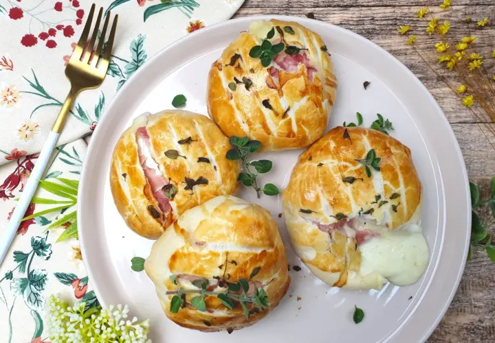 Quattro fagottini dorati con prosciutto e **tomino fuso** che cola, guarniti con erbe aromatiche su un piatto rosa.