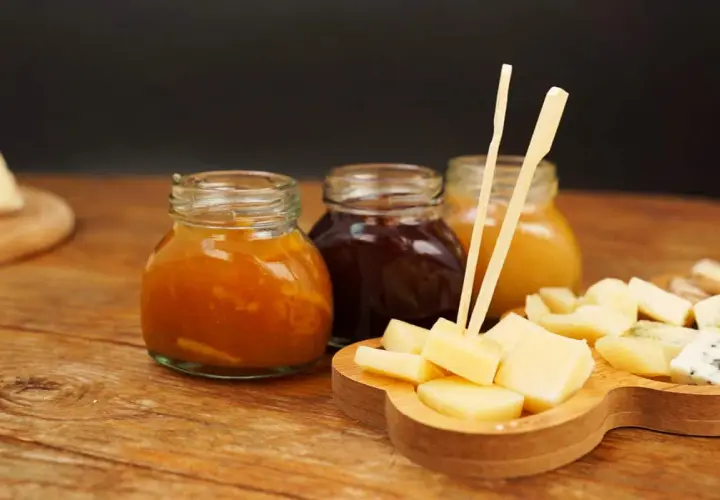 Tre vasetti di marmellata, uno arancione, uno scuro e uno giallo, accompagnano un tagliere di formaggio a cubetti, pronto per essere gustato.