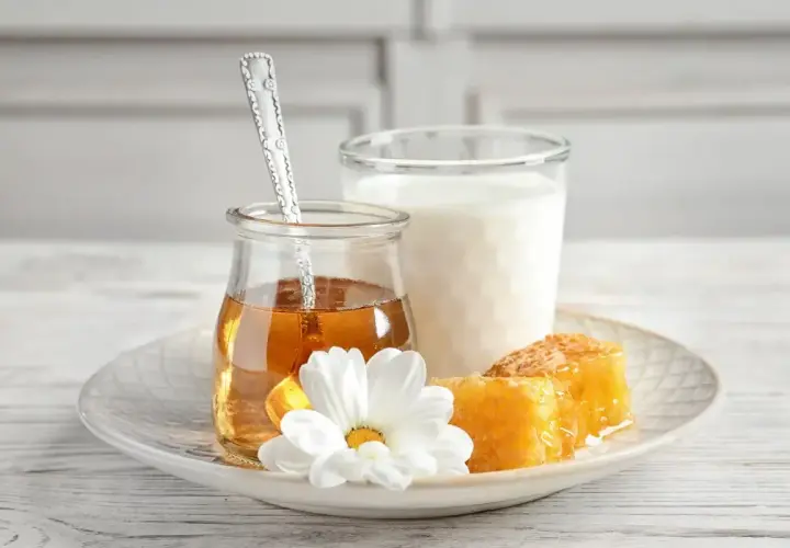 Un bicchiere di latte, un vasetto di miele dorato con un cucchiaino, e favi di miele su un piatto con un fiore bianco. Alcuni credono che latte caldo e miele faccia male.