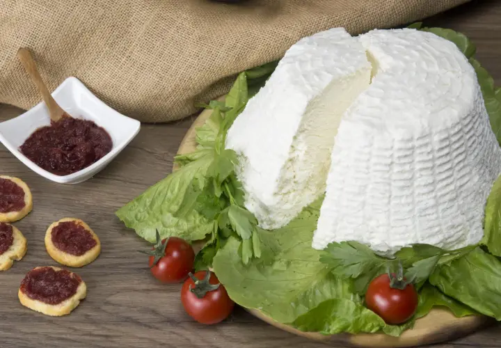 Un grande blocco di ricotta fresca, circondato da lattuga e pomodorini, con bruschette e marmellata in una ciotola. Diversi tipi di ricotta sono presentati in modo invitante.
