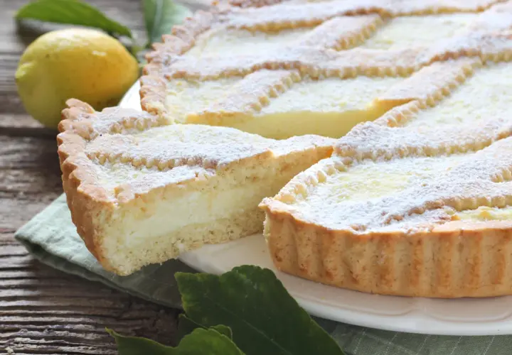 Una fetta di crostata di ricotta, cosparsa di zucchero a velo, accanto a un limone e foglie verdi su un tavolo di legno.