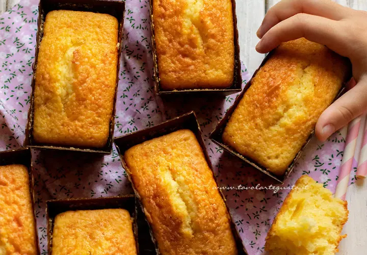 Plumcake yogurt dorati e soffici, pronti per essere gustati. Una mano ne afferra uno, mentre un pezzetto mostra la morbidezza interna.