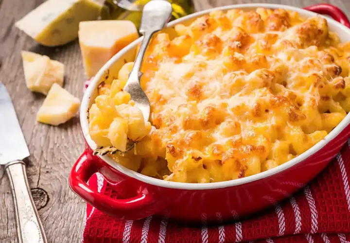 Mac and cheese dorato e filante in una teglia rossa, con pezzi di formaggio e una forchetta pronta a servirlo.