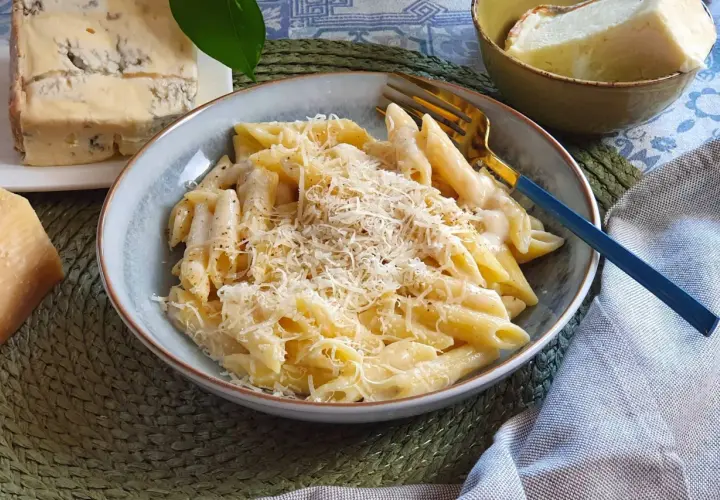 Piatto di penne cremose, condite con una ricca salsa di spaghetti ai 4 formaggi, guarnite con parmigiano grattugiato. Accanto, pezzi di formaggio fresco e stagionato.