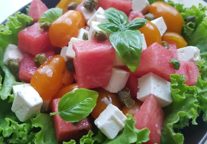 Insalata estiva con cubetti di formaggio di capra, anguria, pomodorini gialli, capperi e basilico fresco su letto di lattuga croccante.
