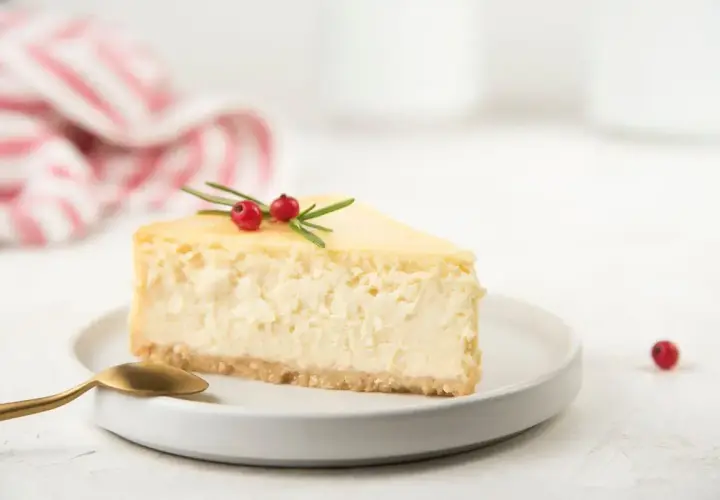 Una fetta di cheesecake con yogurt greco, guarnita con rosmarino e bacche rosse, su un piatto bianco con un cucchiaino dorato.