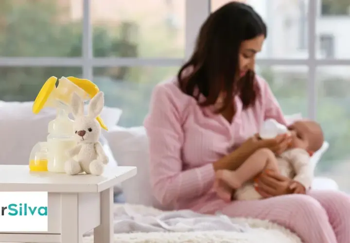 Mamma nutre il neonato con il biberon. Sul tavolino, un coniglietto di peluche e accessori per la pappa, come i latticini e derivati.