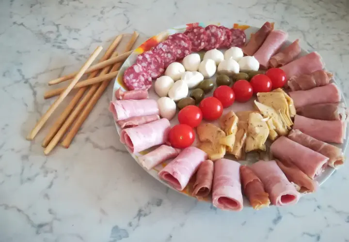 Un'idea per un tagliere di salumi: salame, mozzarelline, olive, pomodorini, carciofini e involtini di prosciutto cotto e crudo, con grissini a lato.