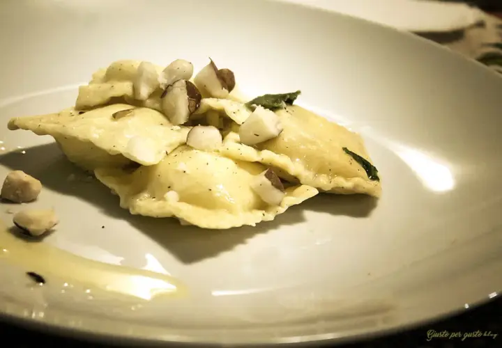 Ravioli pere e pecorino, conditi con nocciole tritate e una foglia di salvia, su un piatto bianco con un filo d'olio.
